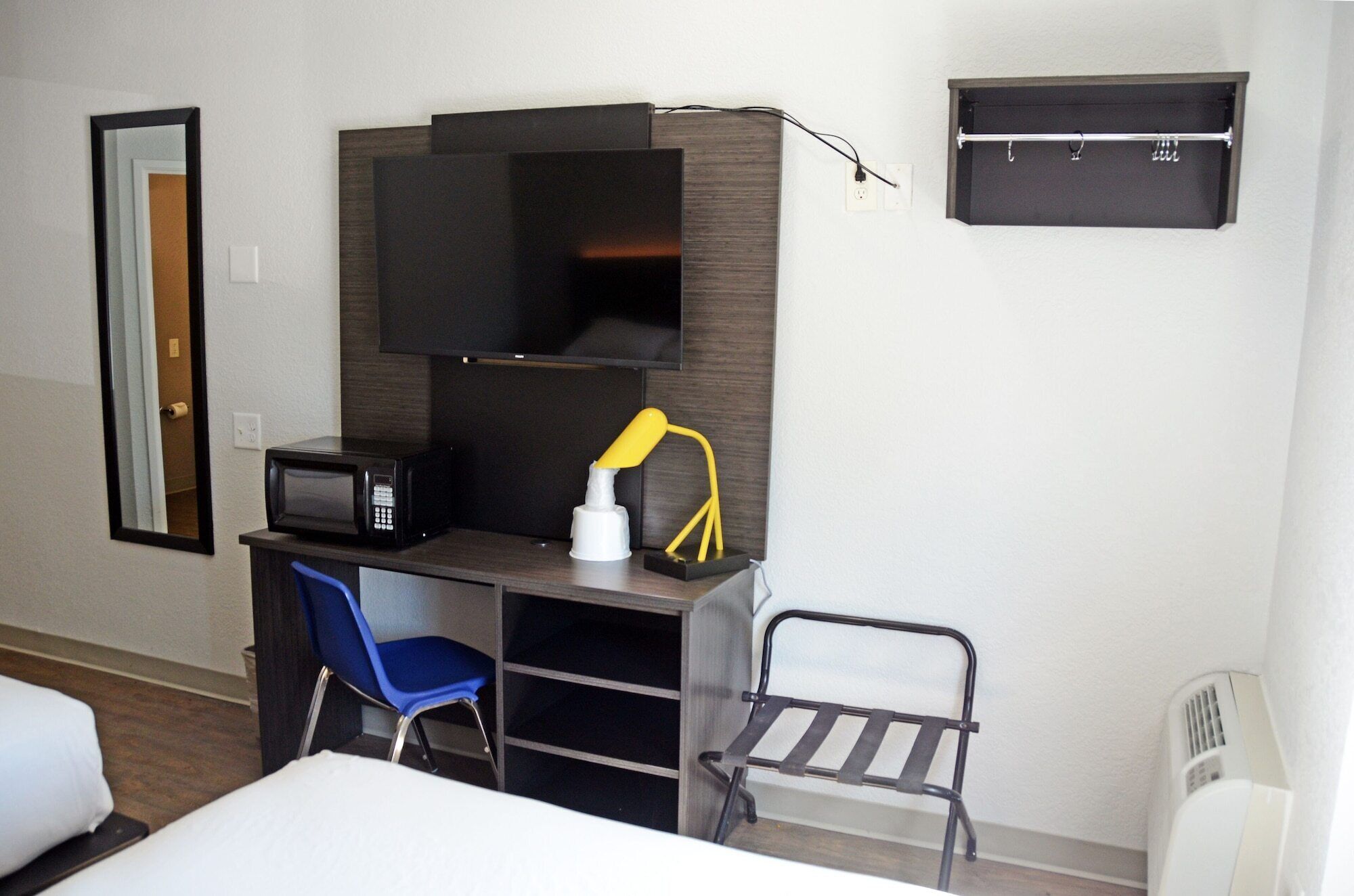 Habitación de hotel pequeña con una cama, un escritorio con silla, microondas, una mesita con lámpara y perchero.