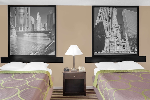 Habitación de hotel con dos camas, una mesita de noche con lámpara y dos cuadros enmarcados con vistas urbanas.