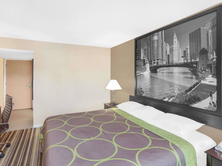 A hotel room with a bed, a desk, a chair, a lamp, and a large cityscape painting on the wall.