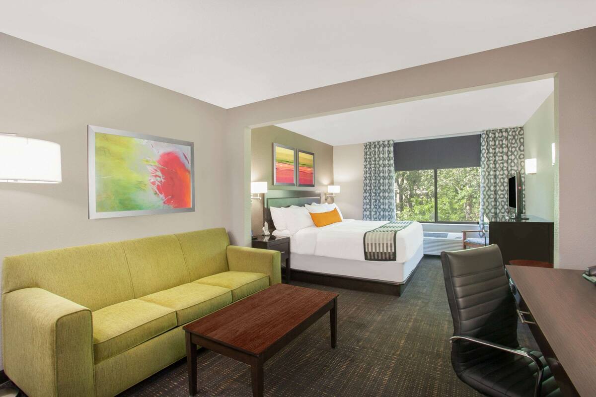 A hotel room with a green sofa, a wooden coffee table, a bed with white bedding and an orange pillow, a desk with a chair, and a large window with curtains.