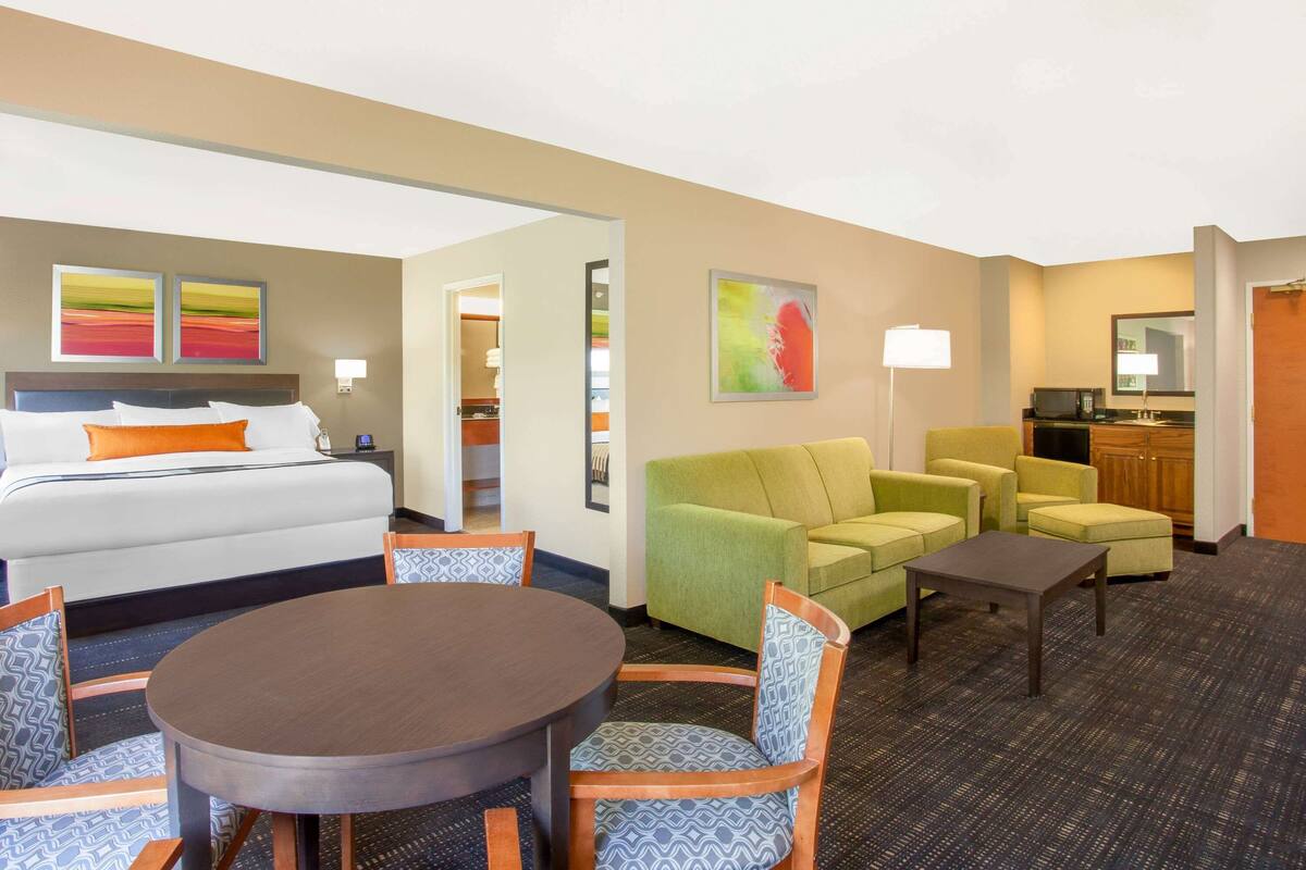 A hotel room with a bed, a dining table with chairs, a sofa set, a small table, and a kitchenette.