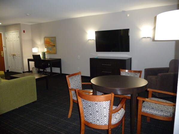 Habitación de hotel con zona de comedor que incluye una mesa redonda y seis sillas, un televisor de pantalla plana en la pared, un escritorio con silla y un cuadro colgado.