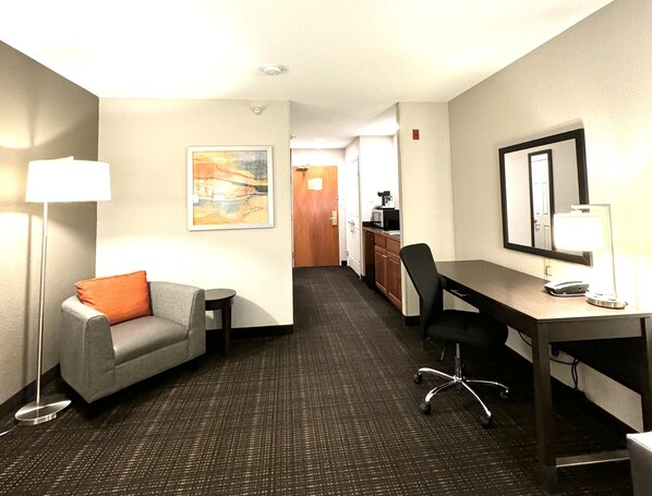 A hotel room with a gray sofa, a desk with a chair, a lamp, a painting on the wall, and a kitchenette in the background.
