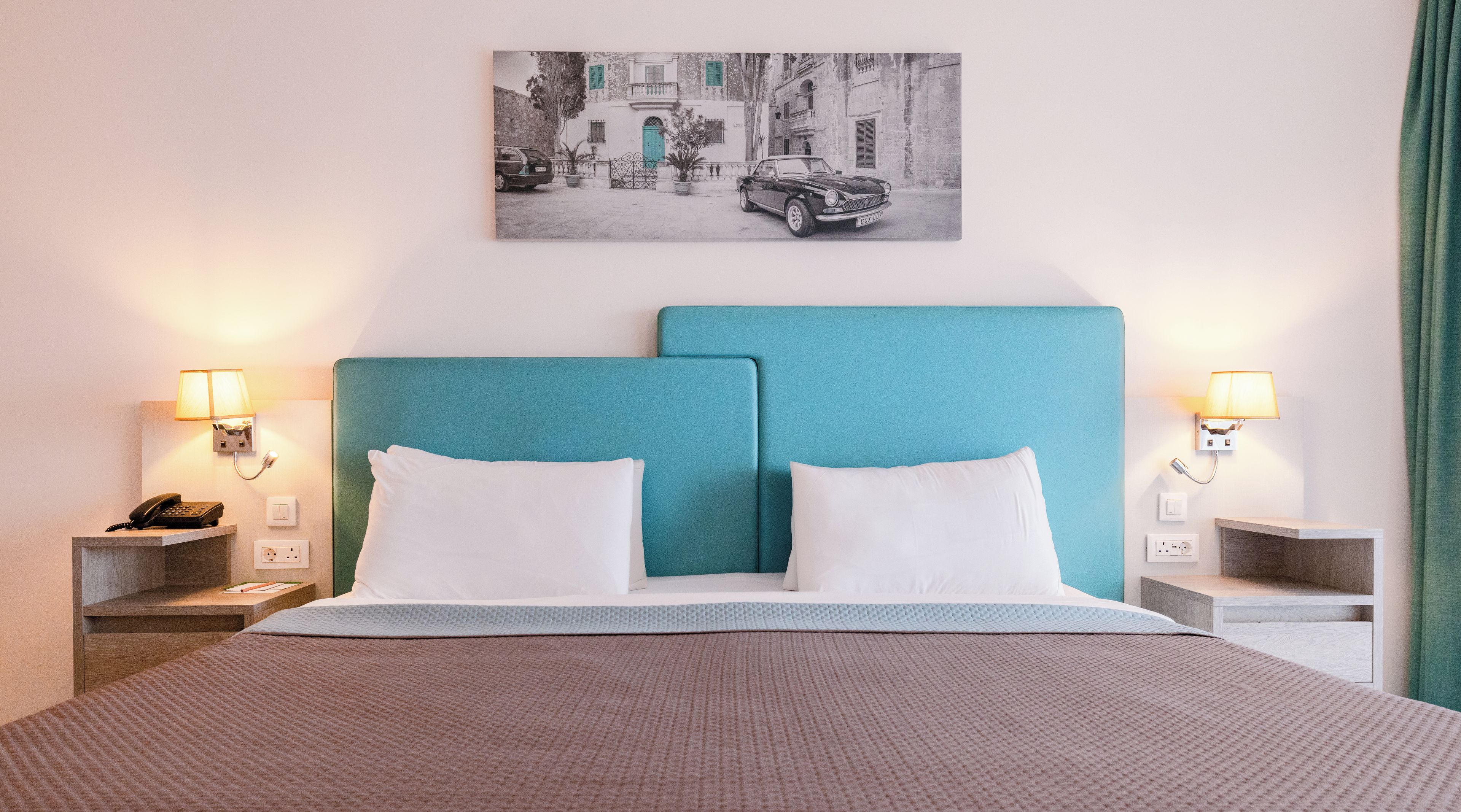 A hotel room with a bed, two bedside tables, a telephone, a lamp, and a wall-mounted picture.