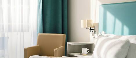 A hotel room with a bed, a chair, a nightstand with a lamp, and a window with curtains.