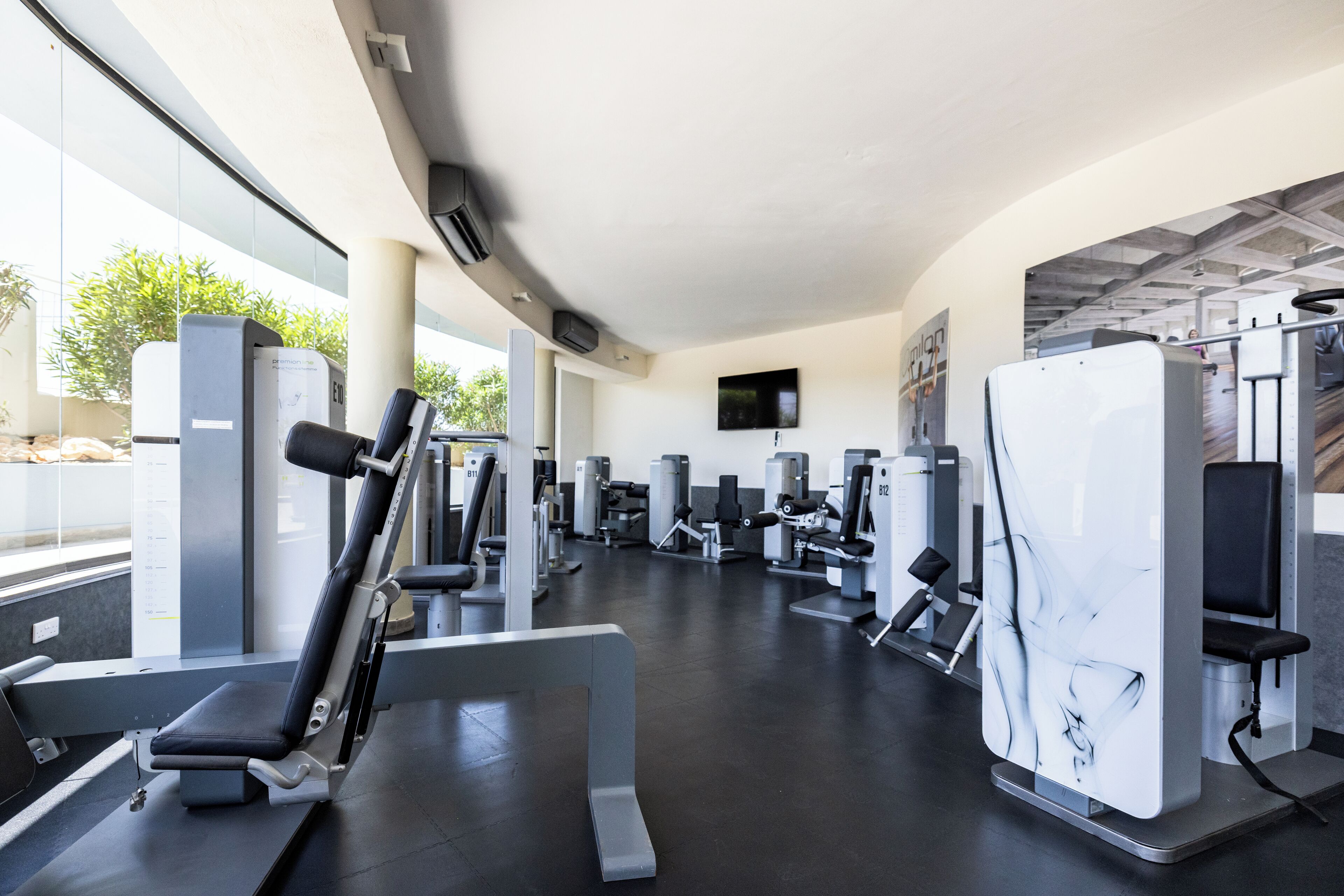 Une salle de sport moderne équipée de divers appareils d’exercice, dotée de grandes fenêtres et d’un plafond plat.