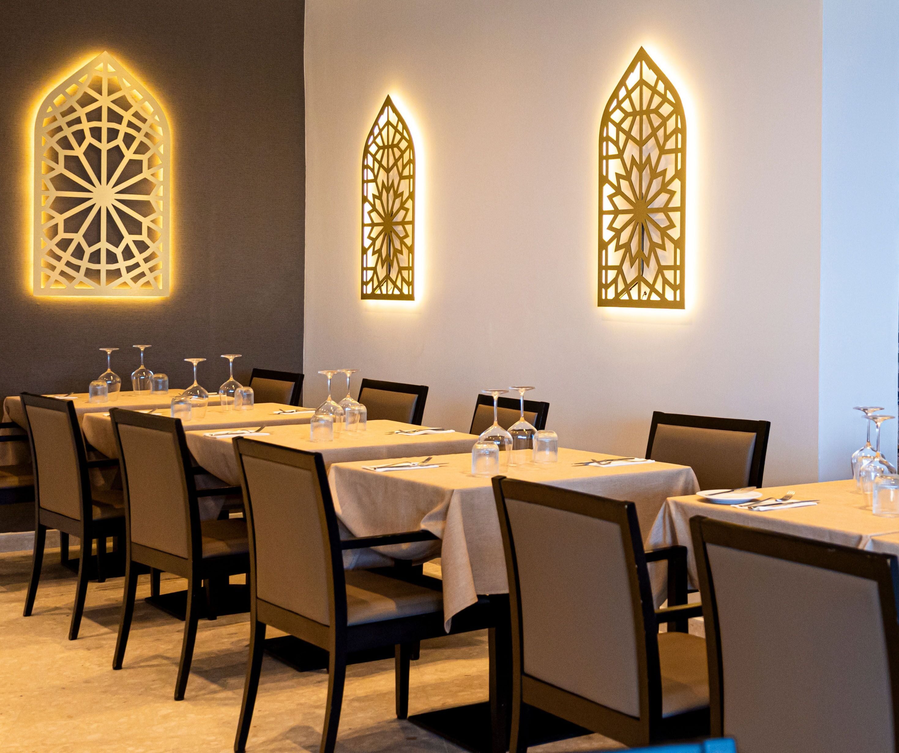 Un espace repas avec des tables dressées pour six personnes, chacune agrémentée de verres à vin et de couverts. Deux panneaux muraux illuminés présentent des motifs complexes.