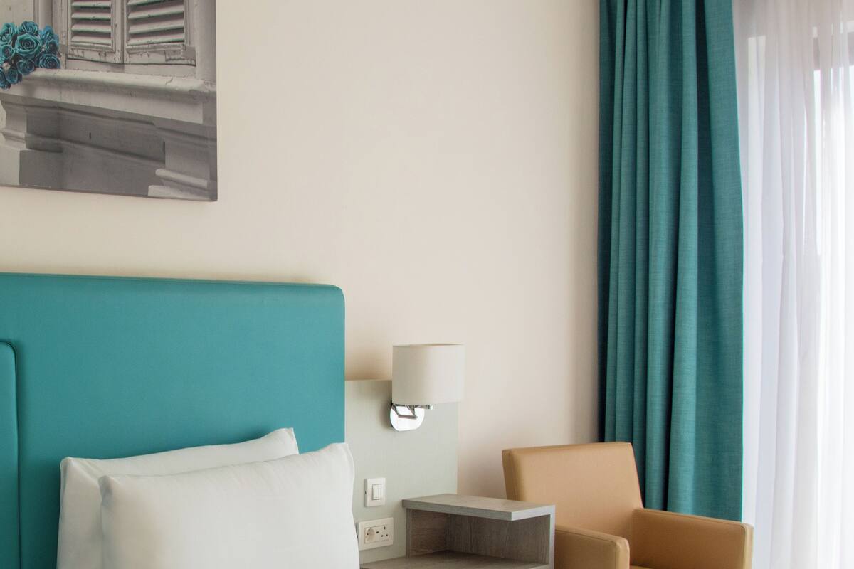 A hotel room with a bed, a nightstand, a lamp, a chair, and a window with curtains.