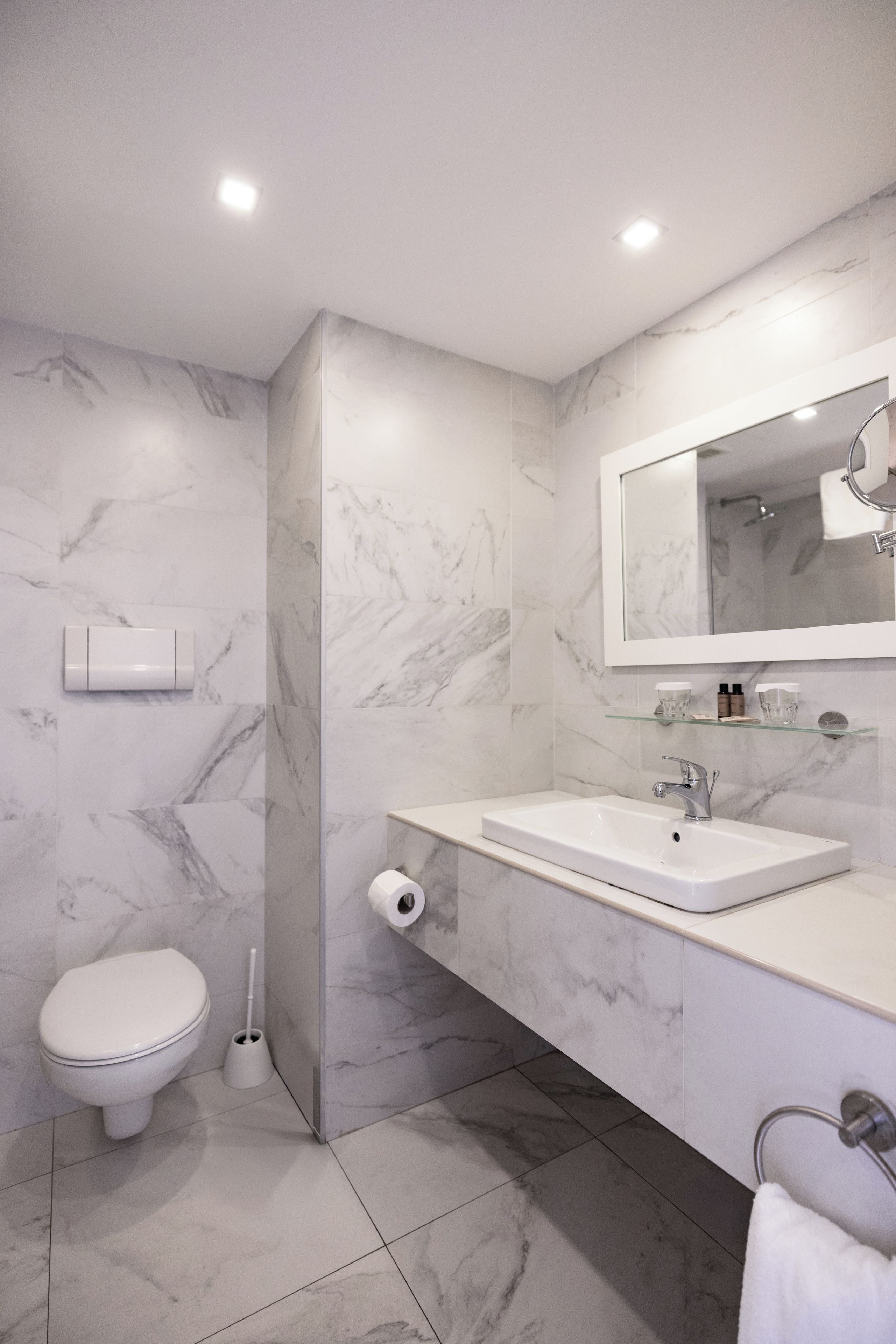 A modern bathroom with a marble wall, a white sink, a toilet, and a glass shelf with toiletries.