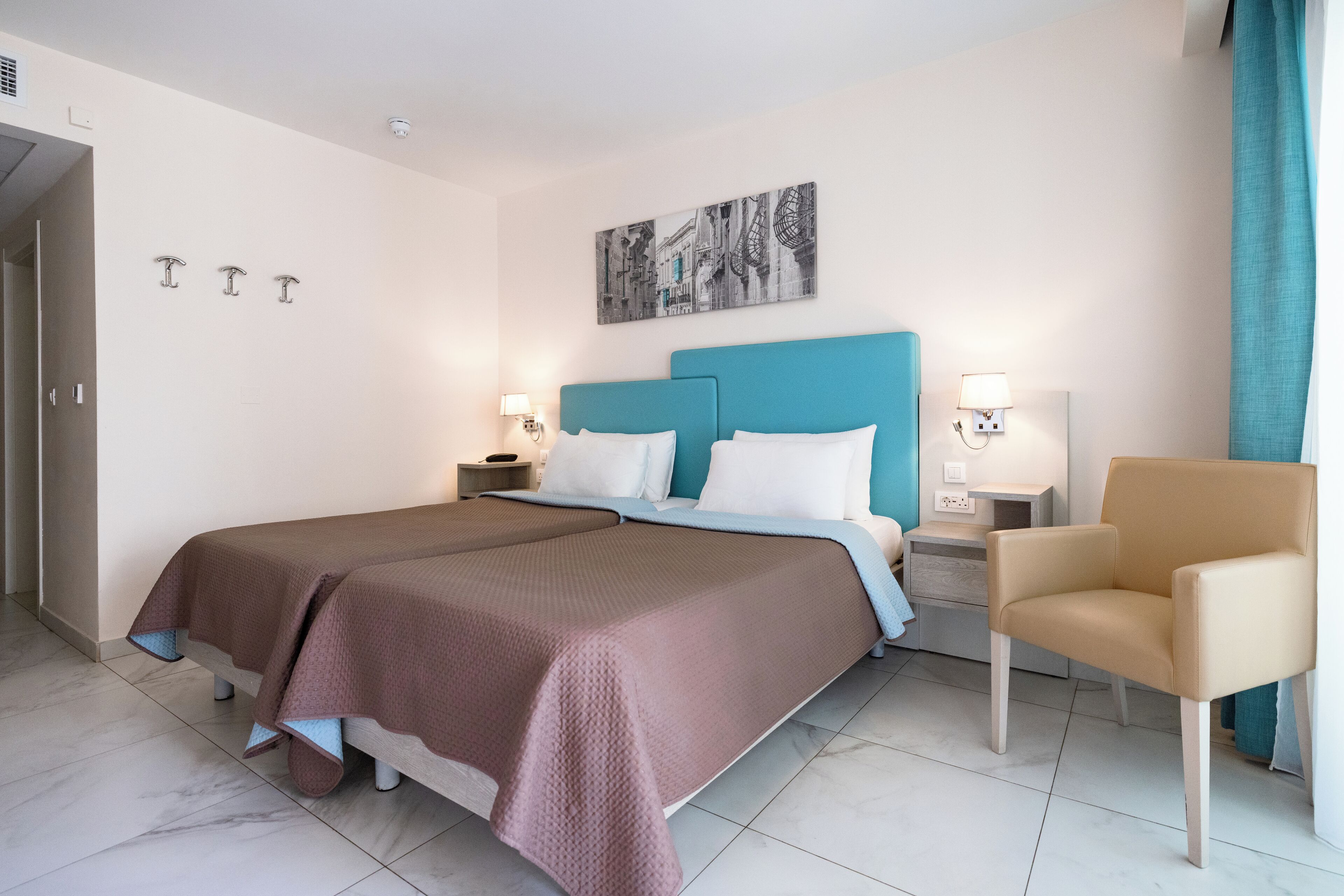 A hotel room with a double bed, a teal headboard, two bedside tables with lamps, a chair, and a wall-mounted artwork.