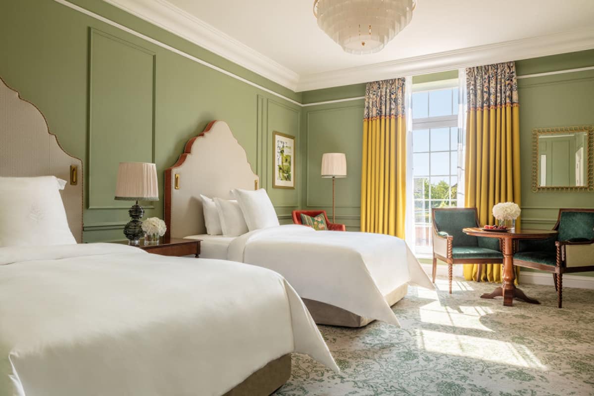 A hotel room with two beds, a sitting area with a chair and table, and a large window with curtains.