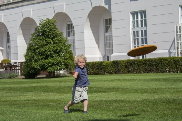 Un niño jugando con un disco volador en una zona verde frente a un edificio con ventanas arqueadas y un gran árbol.