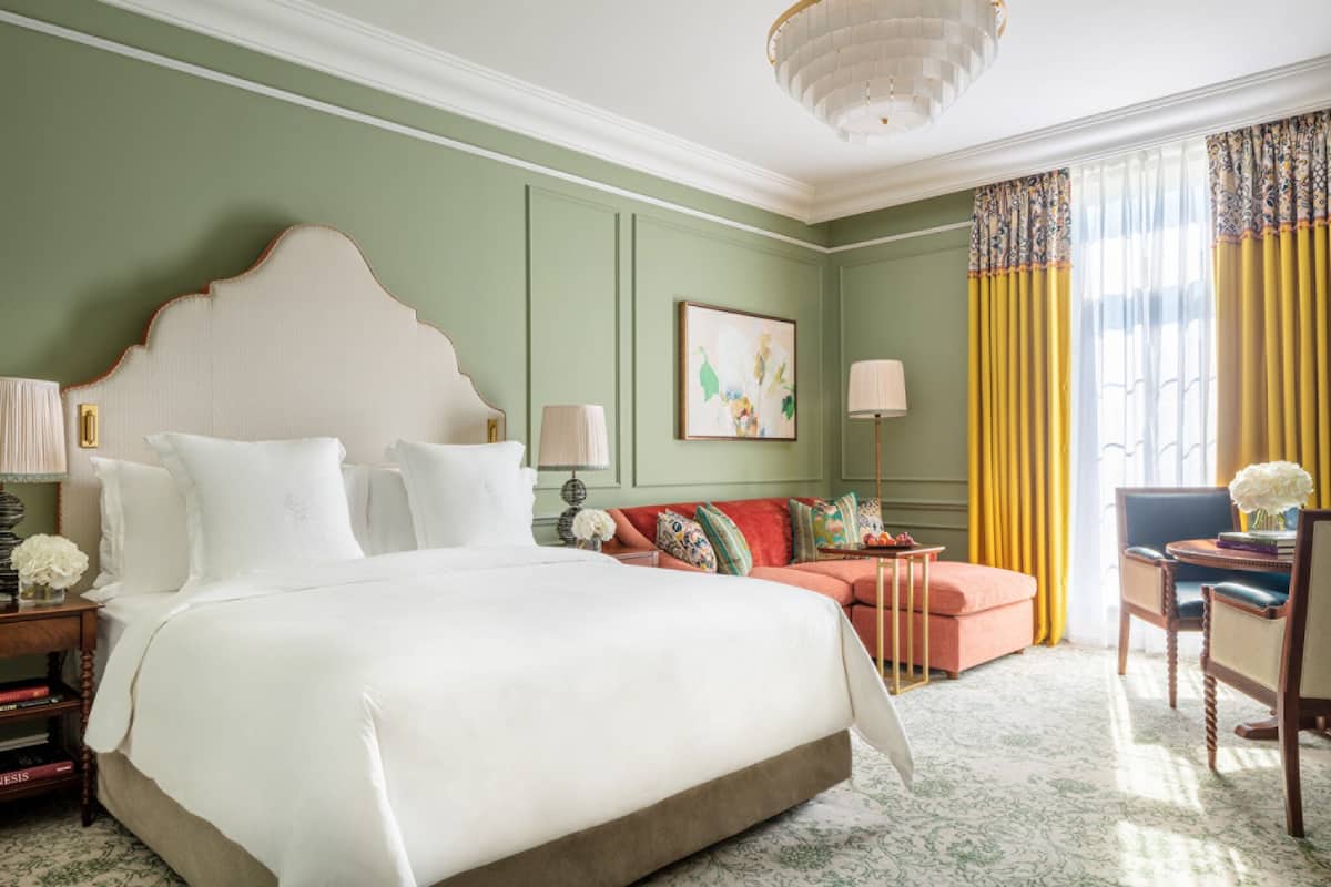 A spacious bedroom with a large bed, a sofa, and a chair. There is a painting on the wall and a window with curtains.