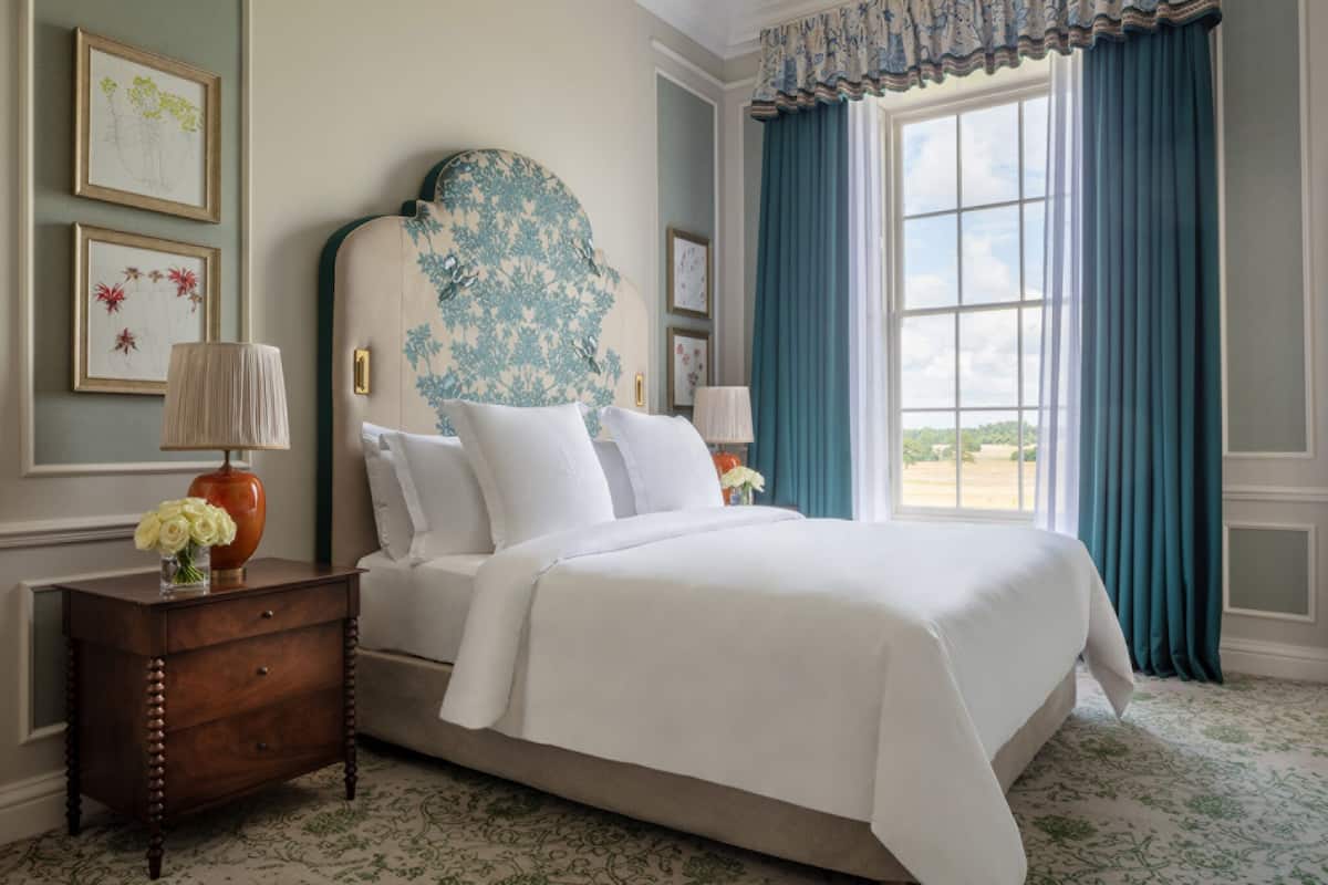 A neatly made bed with white linens, a wooden nightstand with a vase of flowers, and a large window with blue curtains.