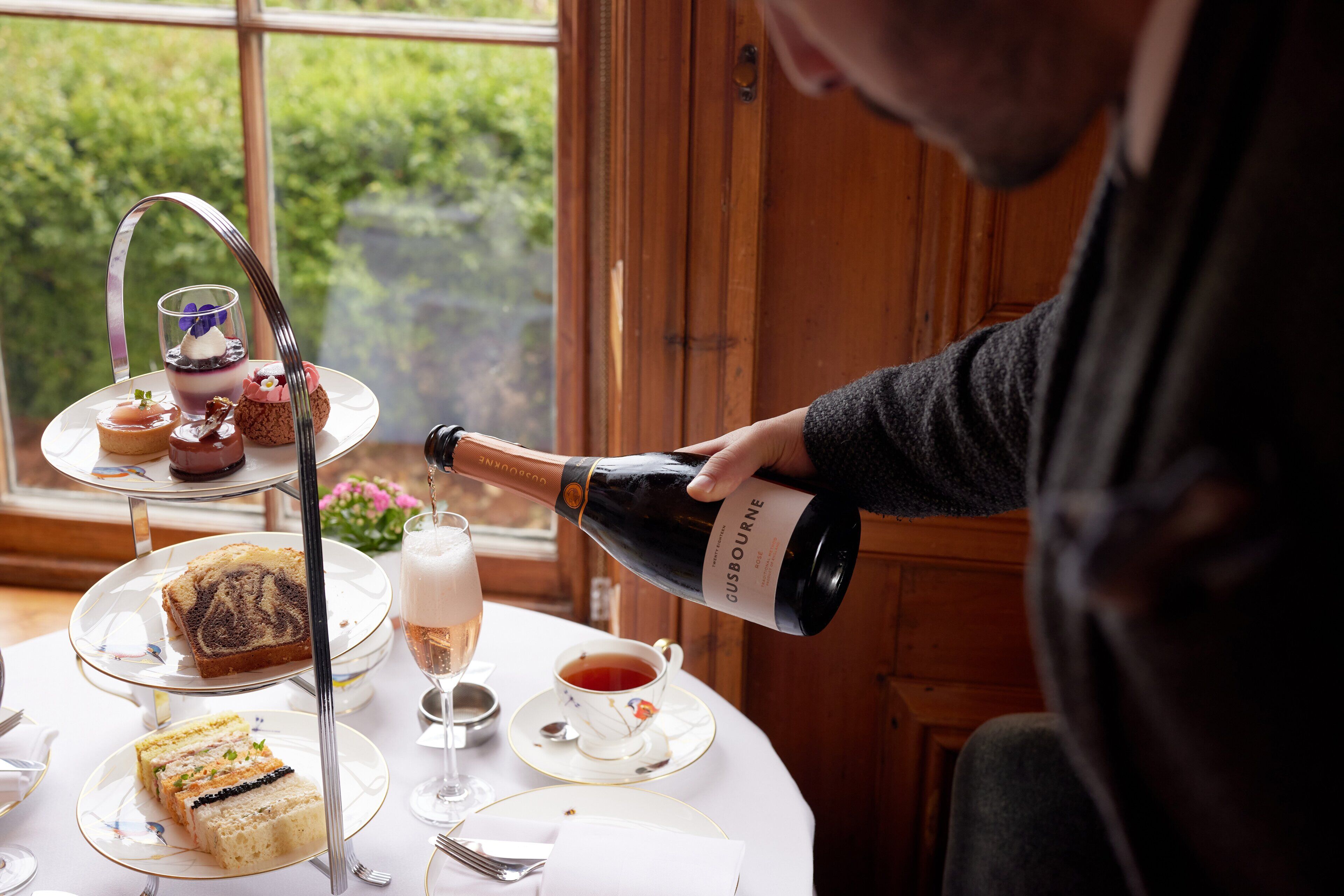 En person häller upp vin från en flaska på ett bord där det står ett våningsfat med desserter och en kopp te.