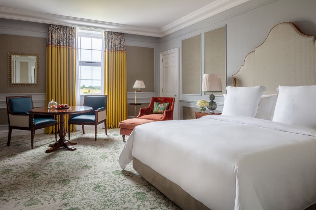 A hotel room with a large bed, a dining table with chairs, a small table with a lamp, and a chair with a cushion.