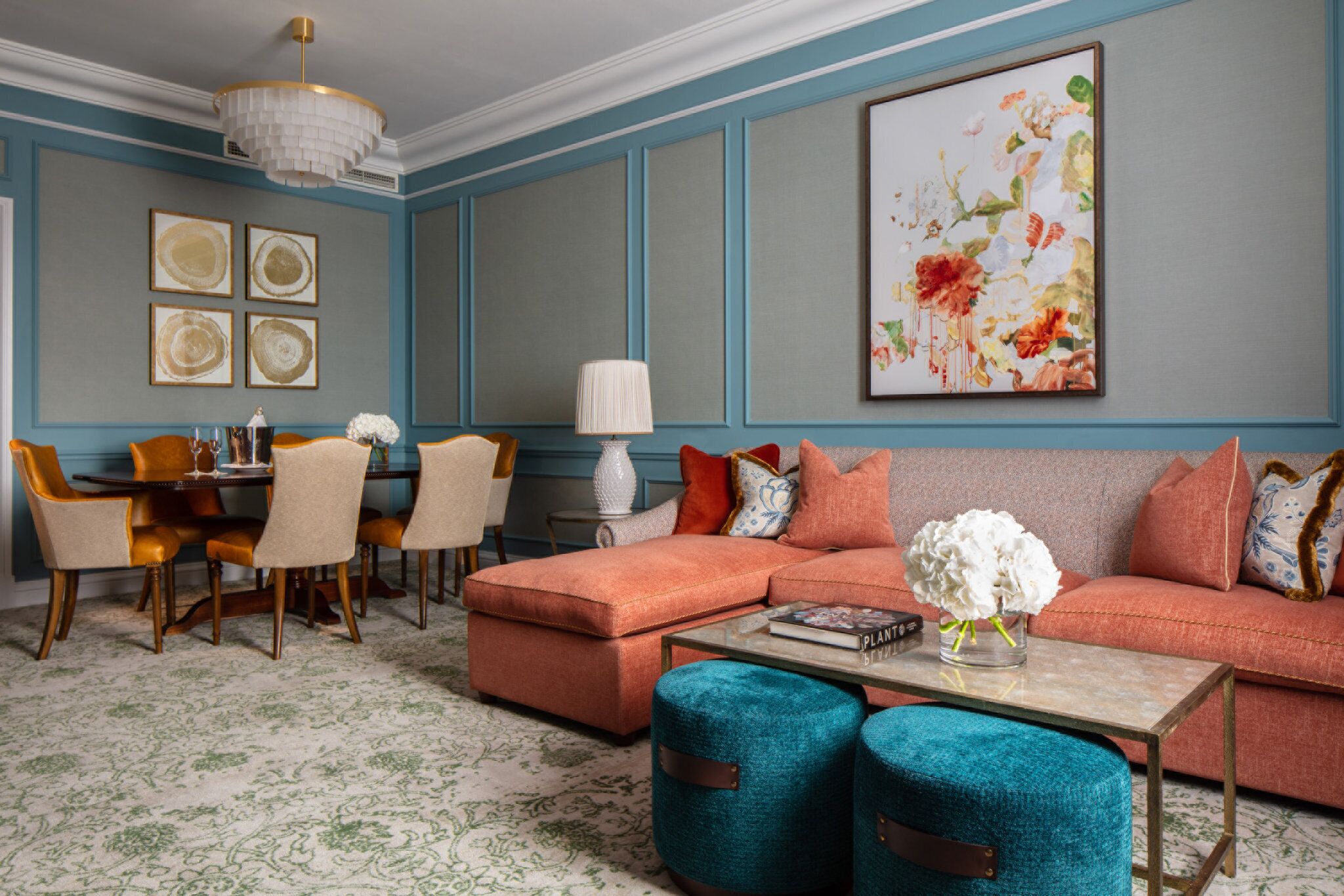A living room with a sectional sofa, ottoman, and teal armchairs. There is a dining table with chairs, a chandelier, and artwork on the walls.