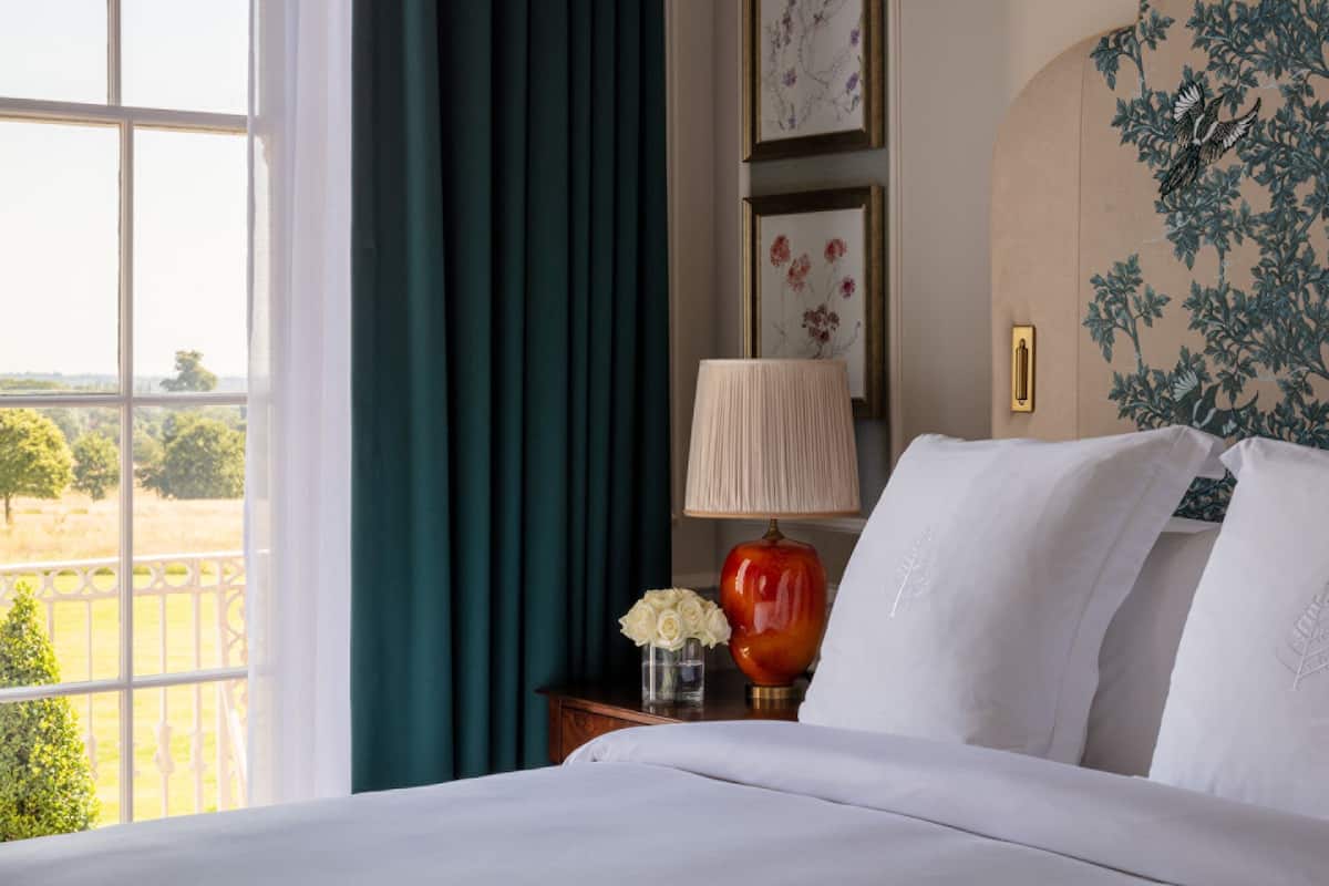 A hotel room with a bed, a nightstand with a lamp, a vase with flowers, and a view of a green landscape through a window.