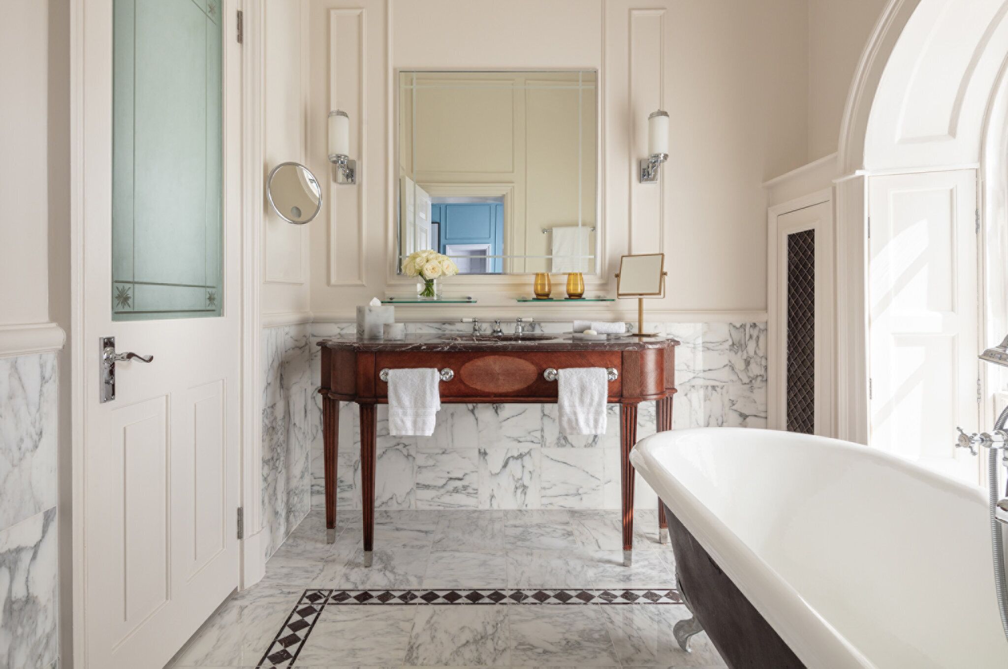 A bathroom with a marble floor, a freestanding bathtub, a vanity with a mirror, and a door with a frosted glass panel.