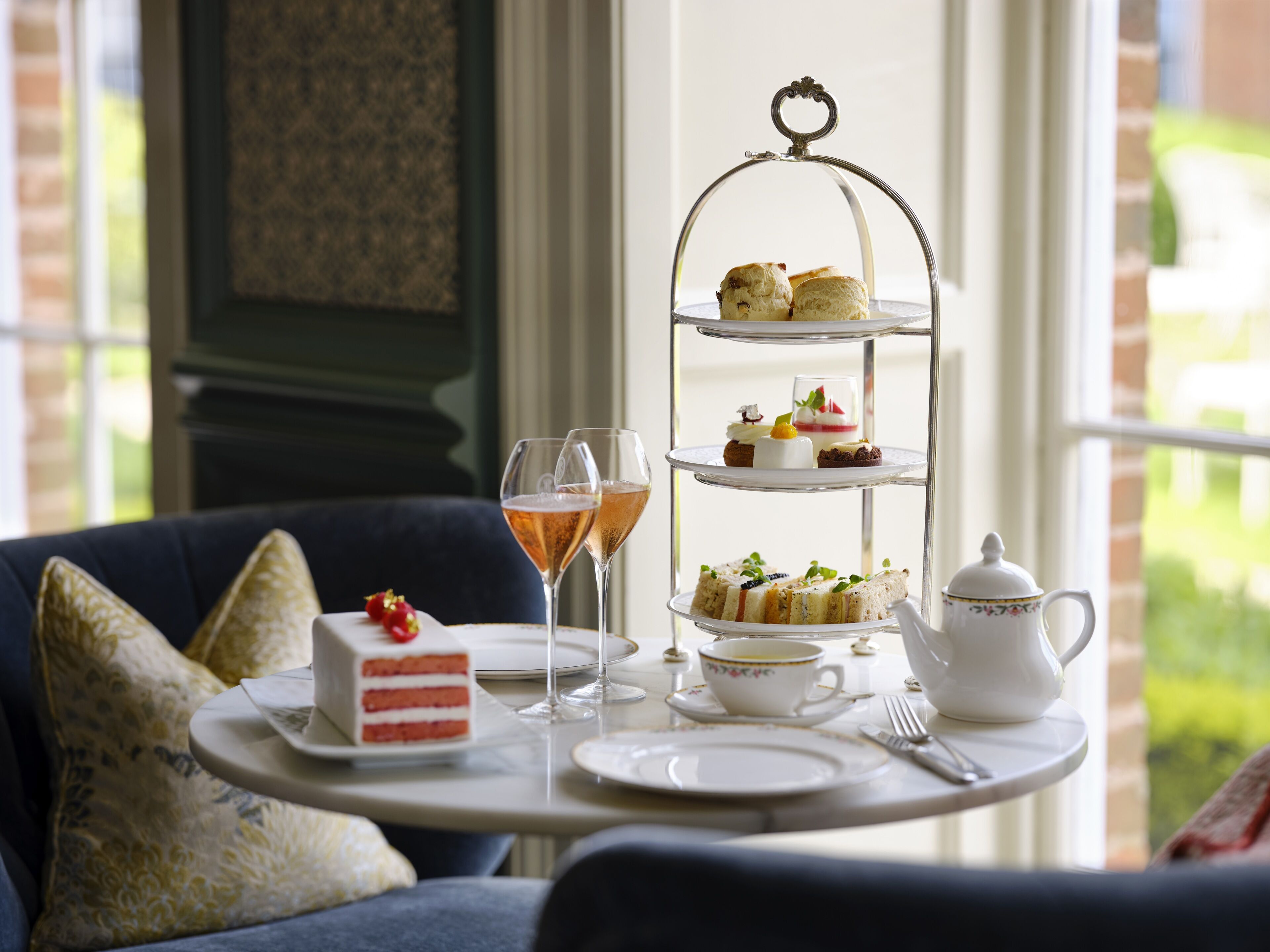 A three-tiered dessert stand with various pastries and a teapot on a table, accompanied by a glass of champagne and a cup of tea.
