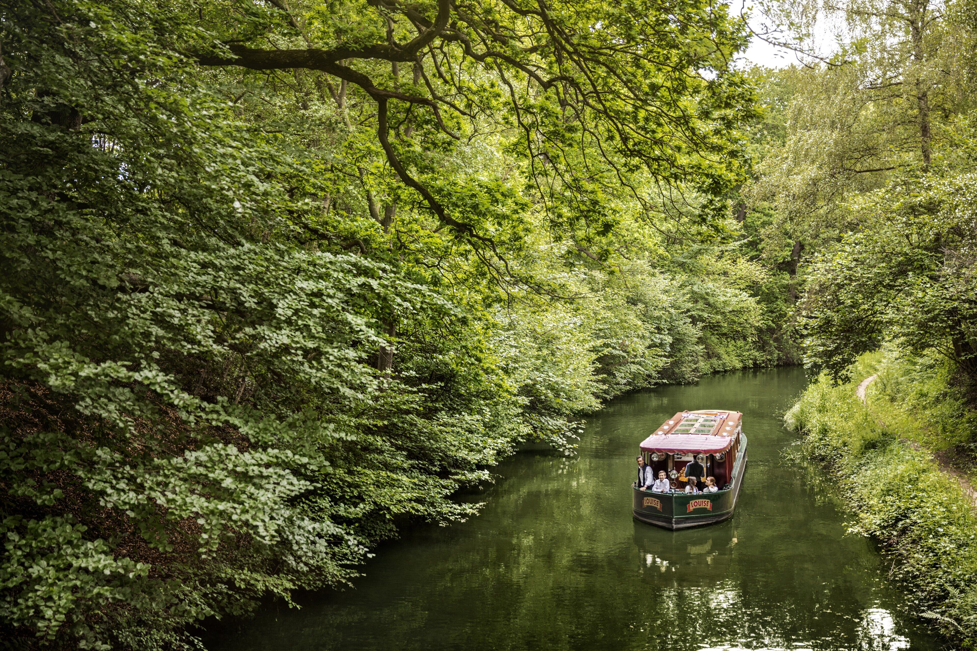 A narrow waterway with dense greenery on both sides. A boat with passengers is navigating the channel.