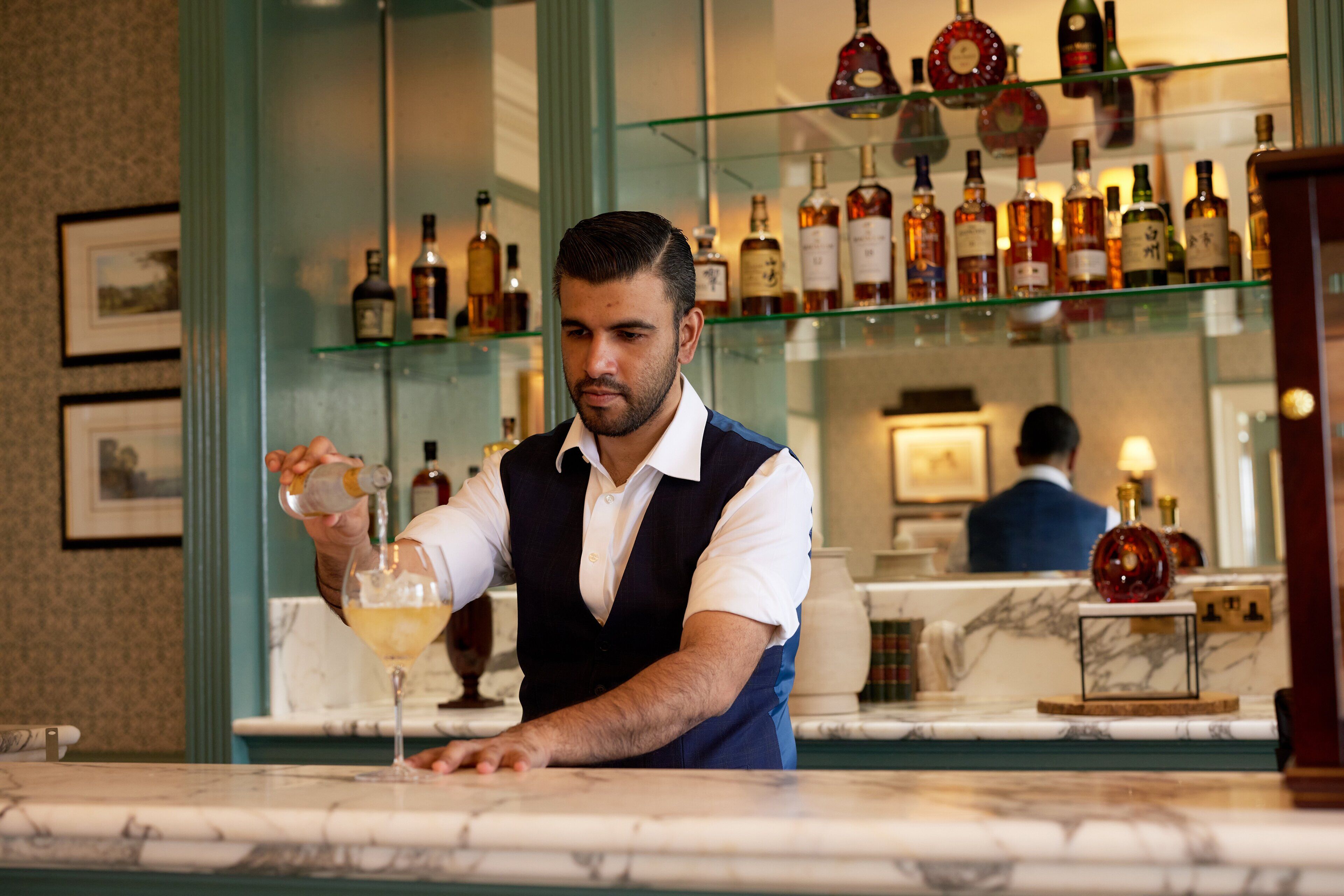 Un bar con un cantinero sirviendo una bebida en un vaso, una barra de mármol y estantes con varias botellas al fondo.