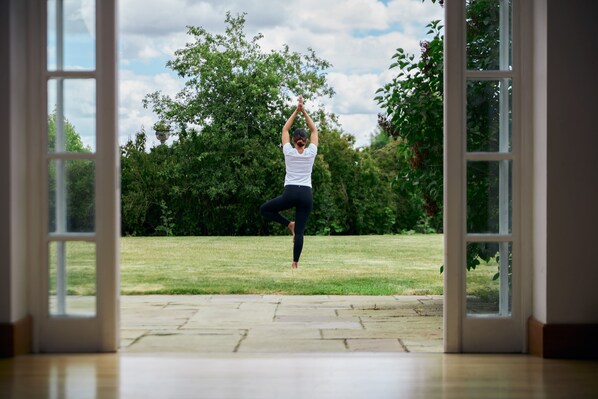 Una persona realiza una postura de yoga en una habitación con amplios ventanales, suelo de madera y vistas a un espacio verde con árboles.