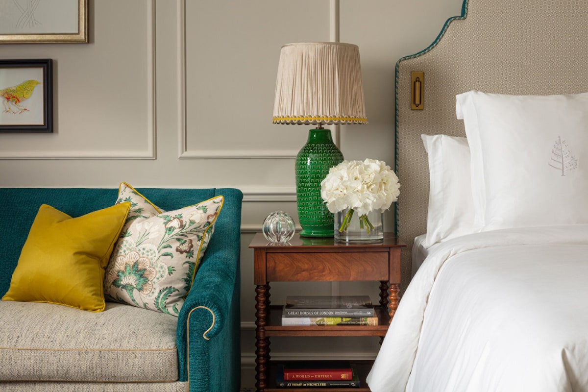 A neatly arranged bedroom with a teal sofa, a green vase with white flowers, and a bedside table with books.
