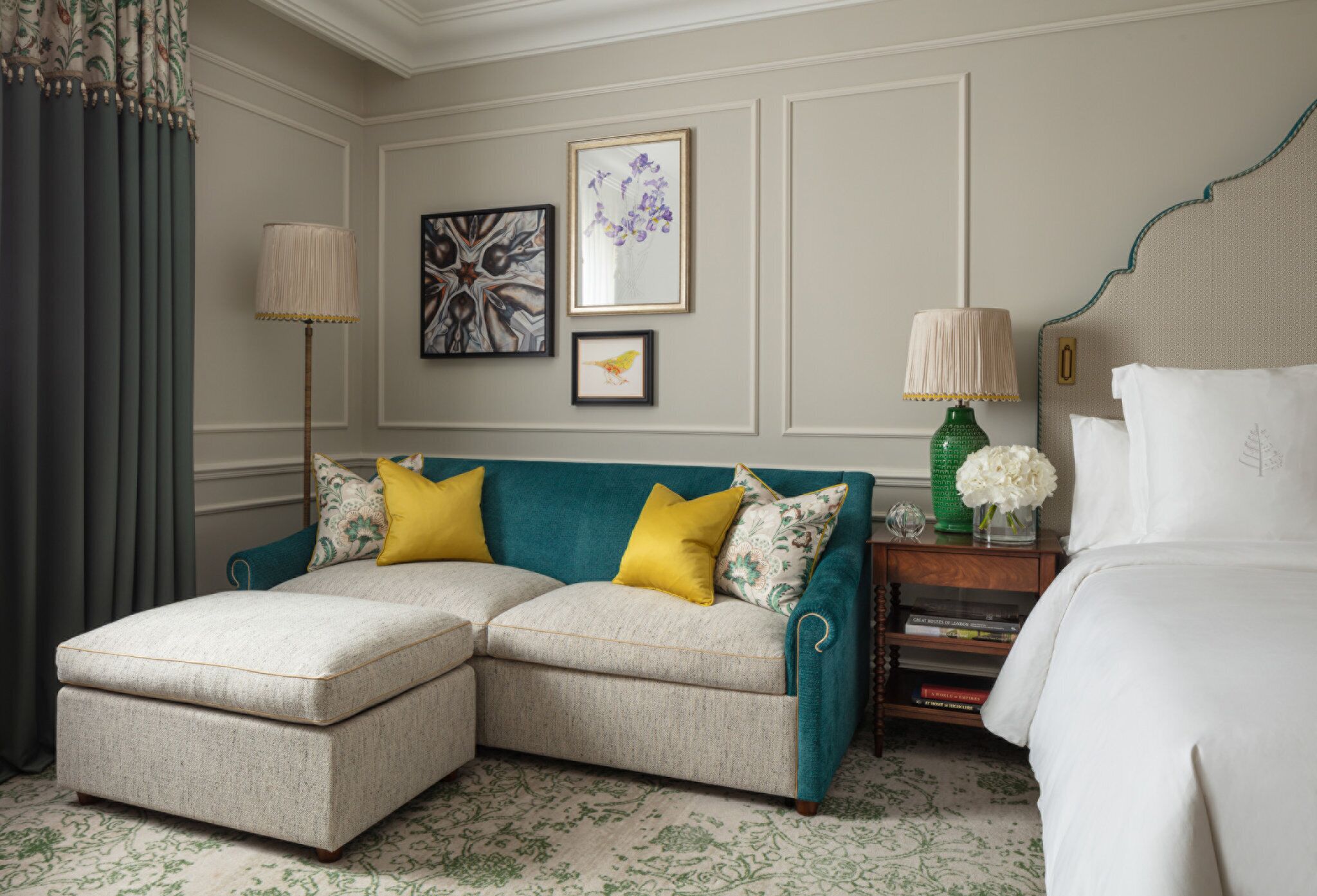 A hotel room with a teal upholstered sofa, a matching ottoman, a bed with white linens, a nightstand with a lamp, and framed artwork on the wall.