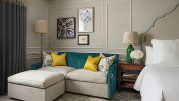 A hotel room with a teal upholstered sofa, a matching ottoman, a bed with white linens, a nightstand with a lamp, and framed artwork on the wall.