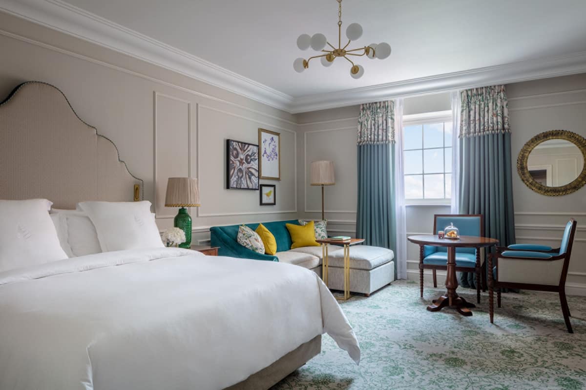 A hotel room with a large bed, a sofa, a small table, and a chair. There is a window with curtains and a mirror on the wall.