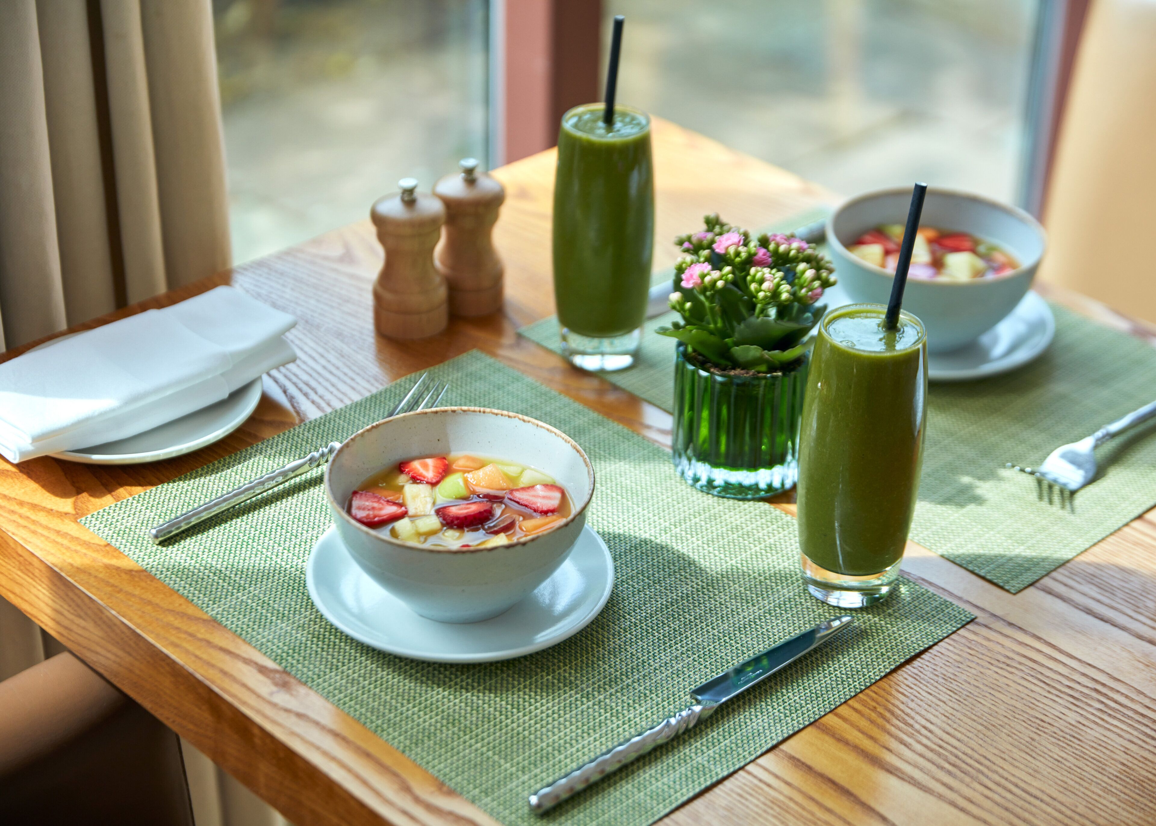 A table with a bowl of fruit salad, two green drinks, a small potted plant, and a salt and pepper shaker.