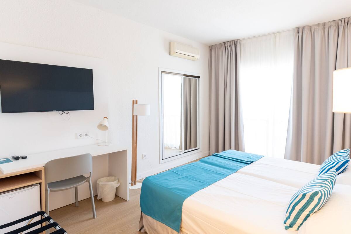 A hotel room with a bed, a desk, a chair, a TV, a window with curtains, and a trash bin.