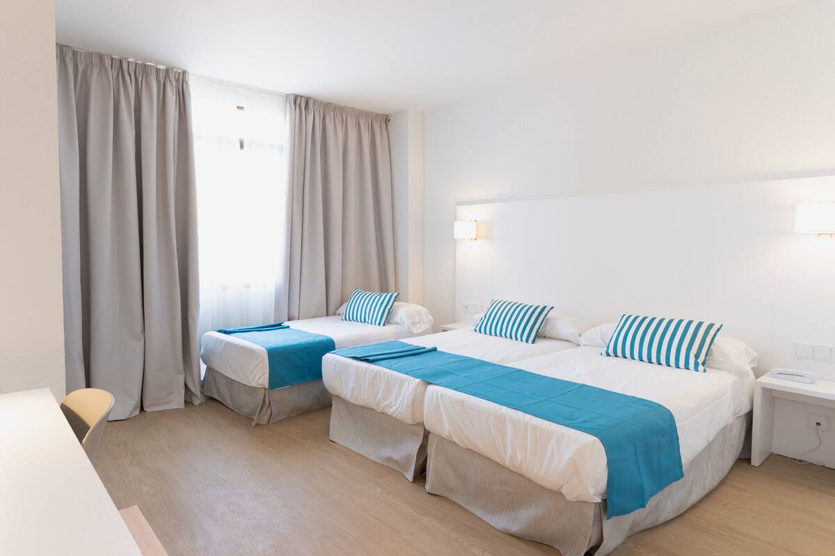 A hotel room with two beds, a desk, a lamp, and a window with curtains.