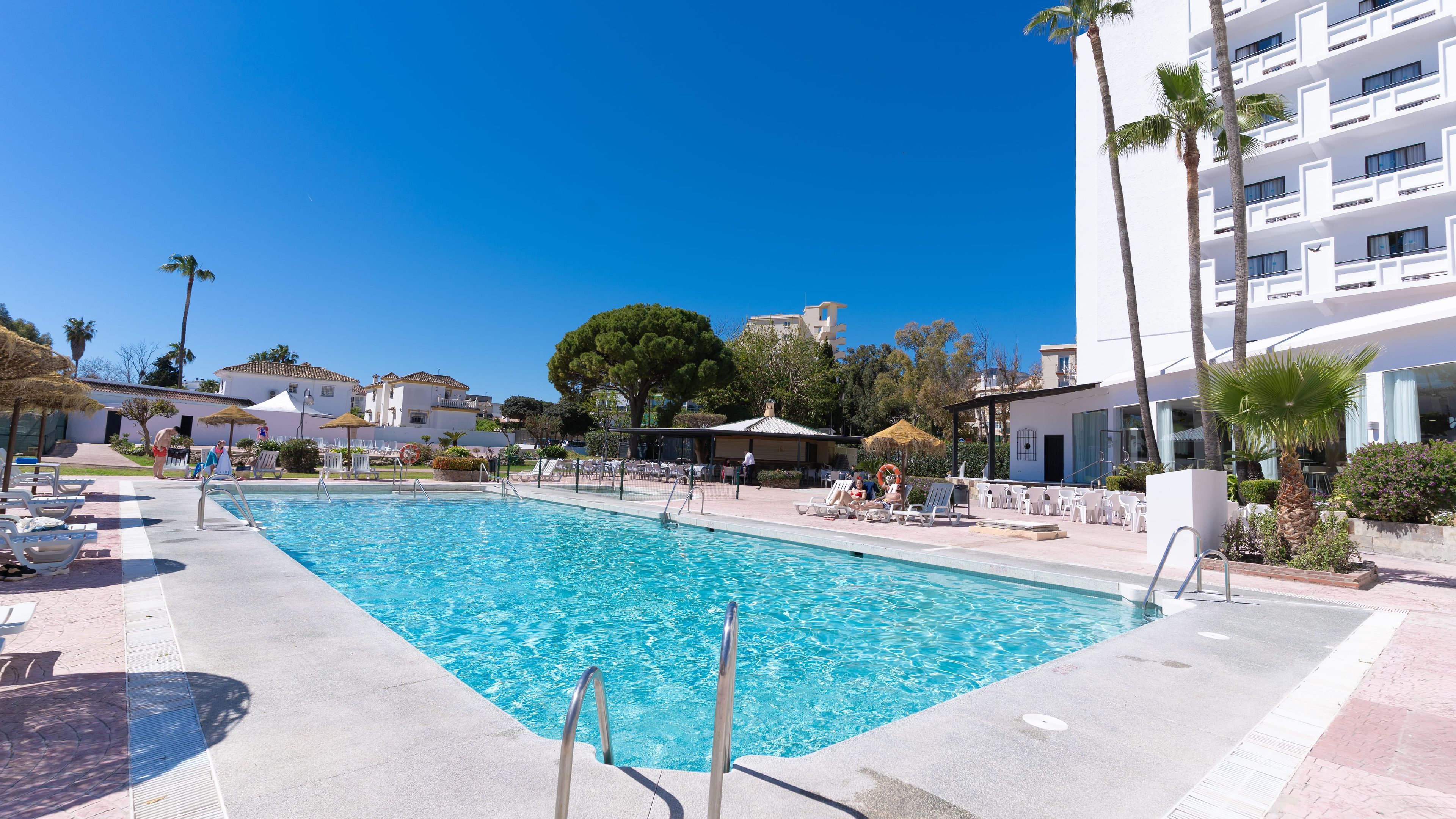 Una piscina con sillas de descanso y sombrillas, rodeada de edificios y palmeras.