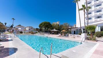Una piscina con sillas de descanso y sombrillas, rodeada de edificios y palmeras.