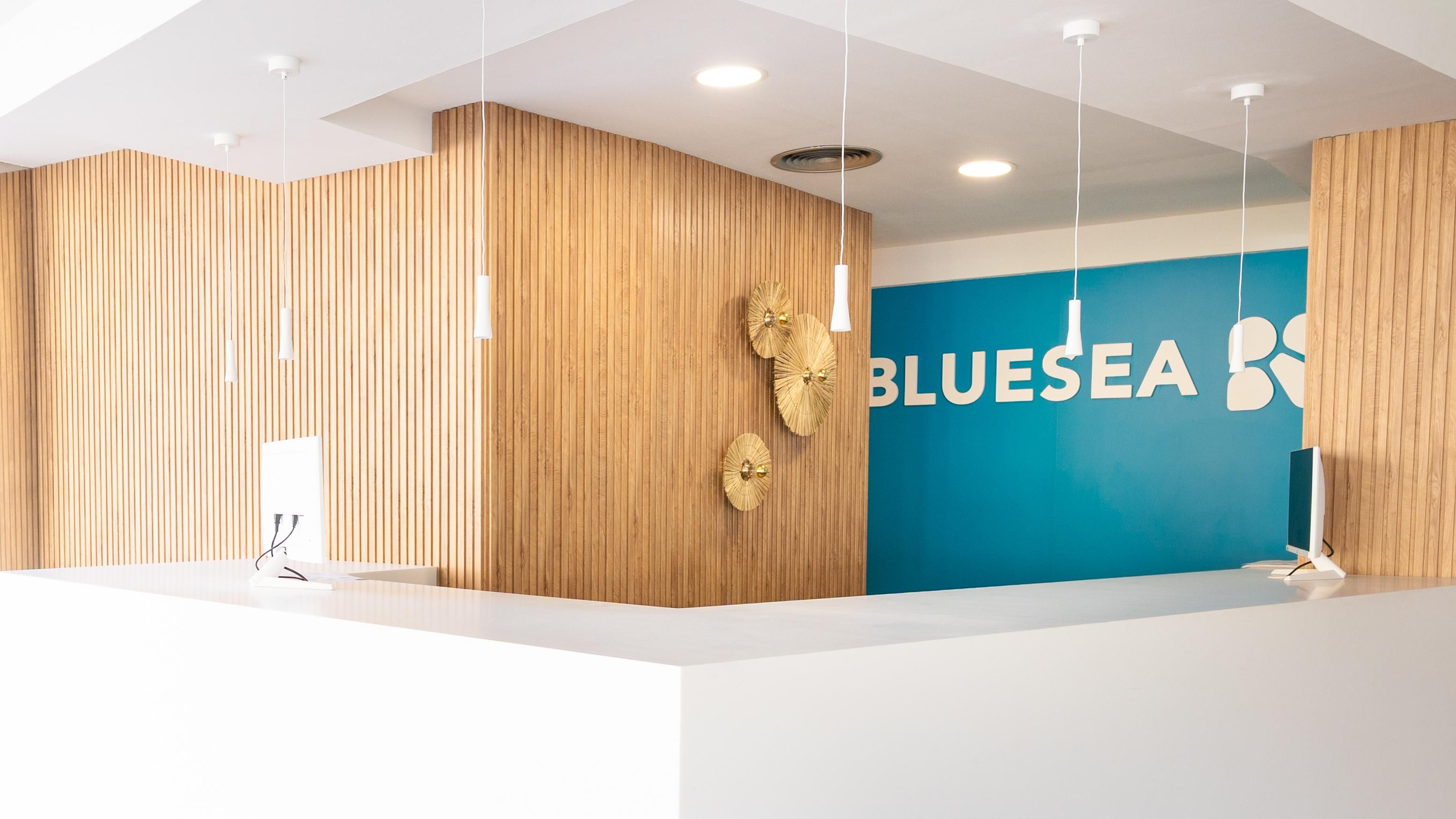 Un comptoir d’accueil avec un mur en panneaux de bois et un mur bleu sur lequel figure le mot « BLUESEA » et un logo. Des luminaires suspendus sont placés au-dessus du comptoir.