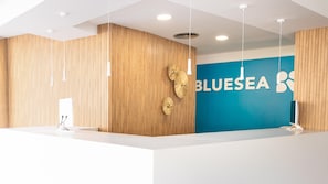 Una recepción con un panel de madera en la pared y una pared azul con la palabra 'BLUESEA' y un logotipo. Hay luces colgantes sobre el mostrador.
