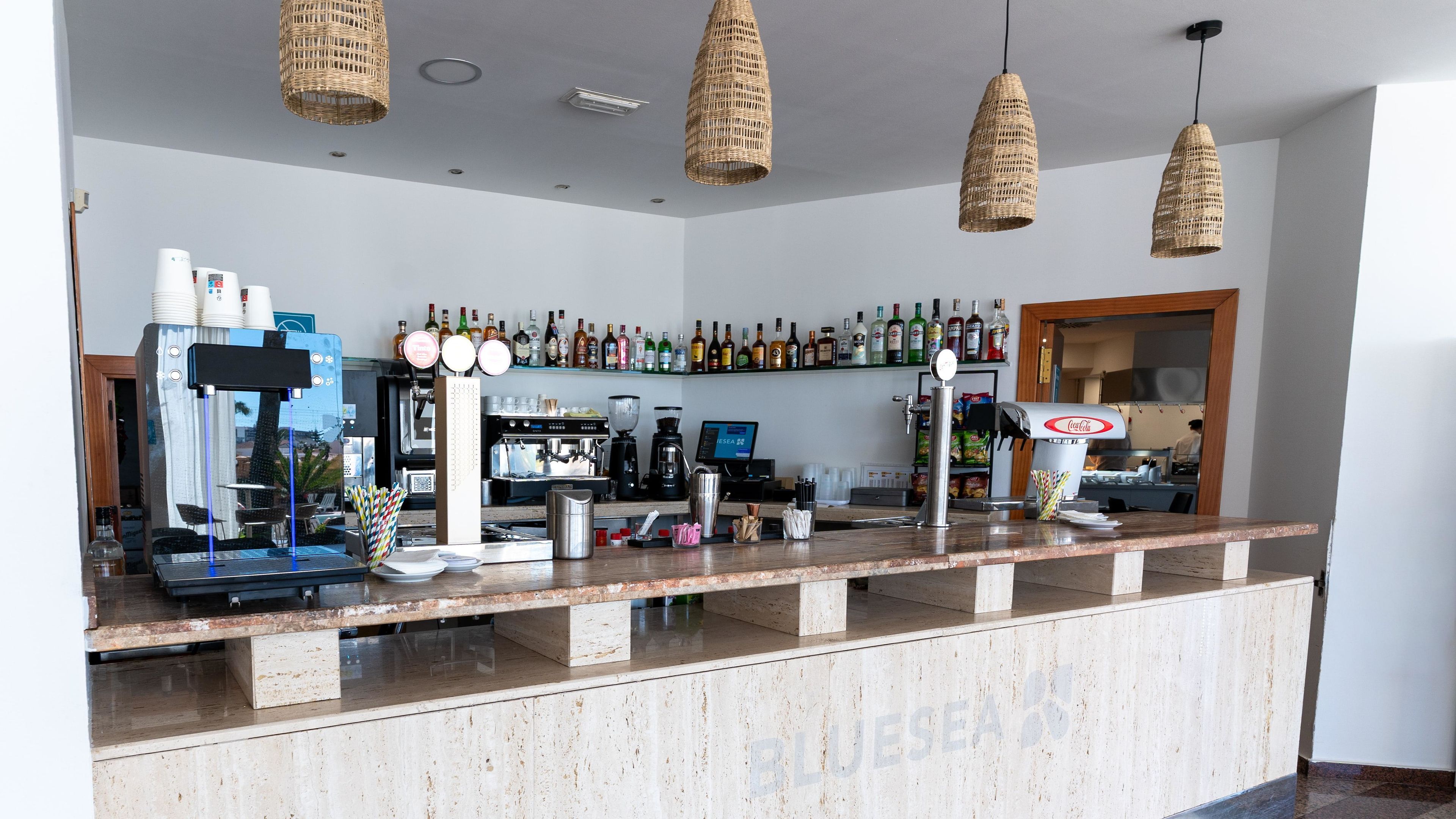 Un espace bar équipé d’une machine à café, d’étagères remplies de bouteilles d’alcool et de luminaires suspendus en osier.