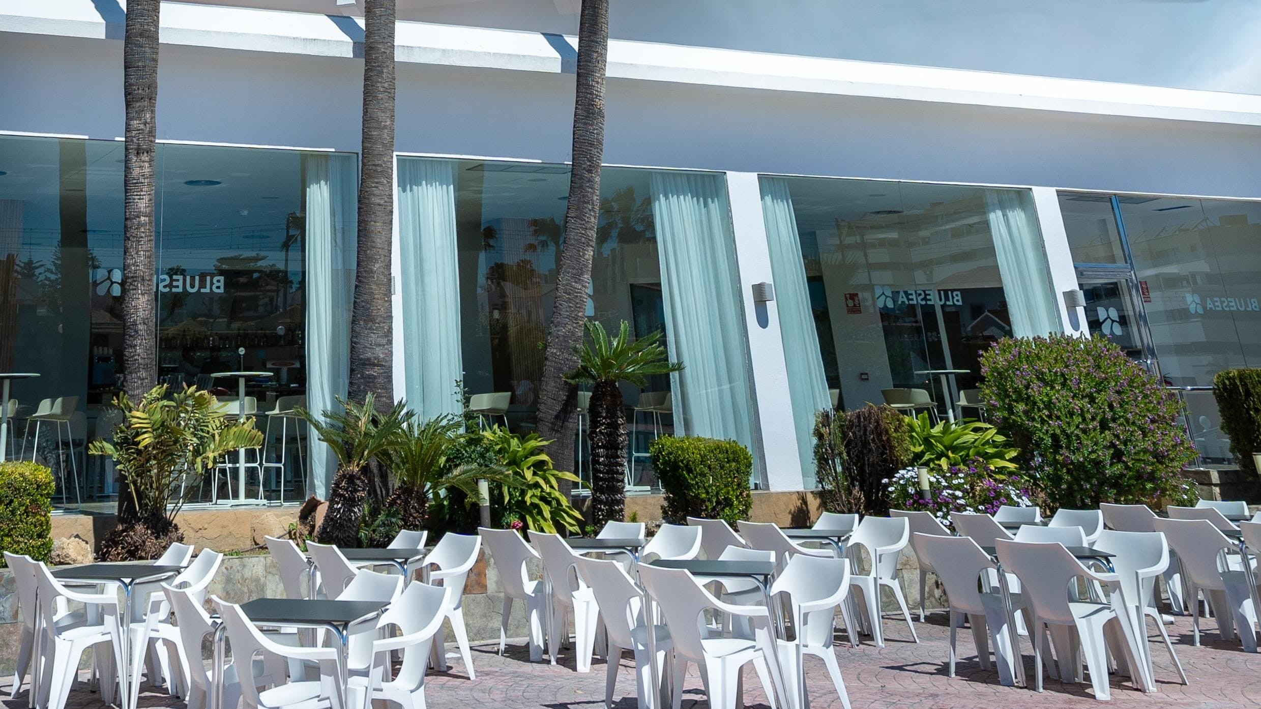 Un espace extérieur aménagé avec des chaises et des tables en plastique blanc, des palmiers et un bâtiment doté de grandes fenêtres en verre et de rideaux.