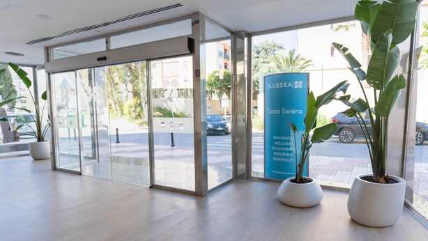 Entrada de hotel com portas de vidro, uma placa azul e plantas em vasos.