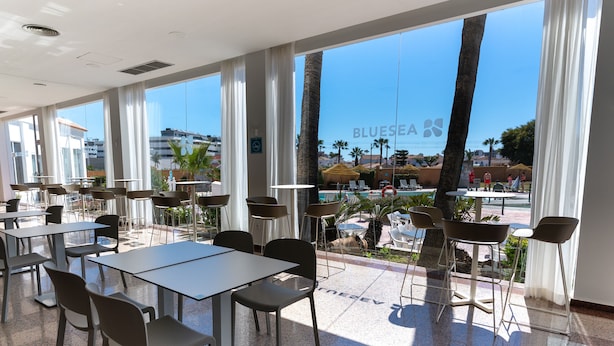 A spacious indoor area with large windows, tables, chairs, and a view of a pool and palm trees. - BLUESEA Costa Serena (Torremolinos)