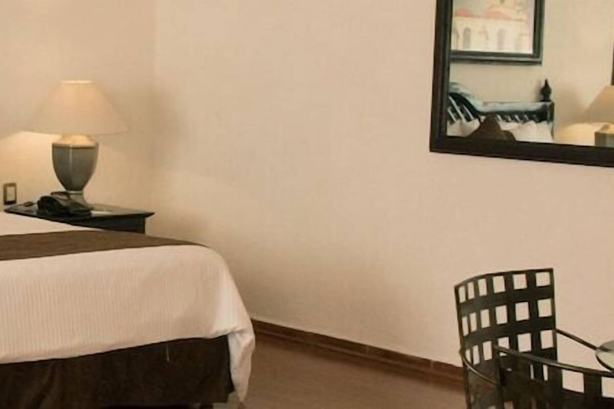 Ein Hotelzimmer mit einem Bett, zwei Nachttischen, einem Sessel und einem an der Wand befestigten Spiegel.