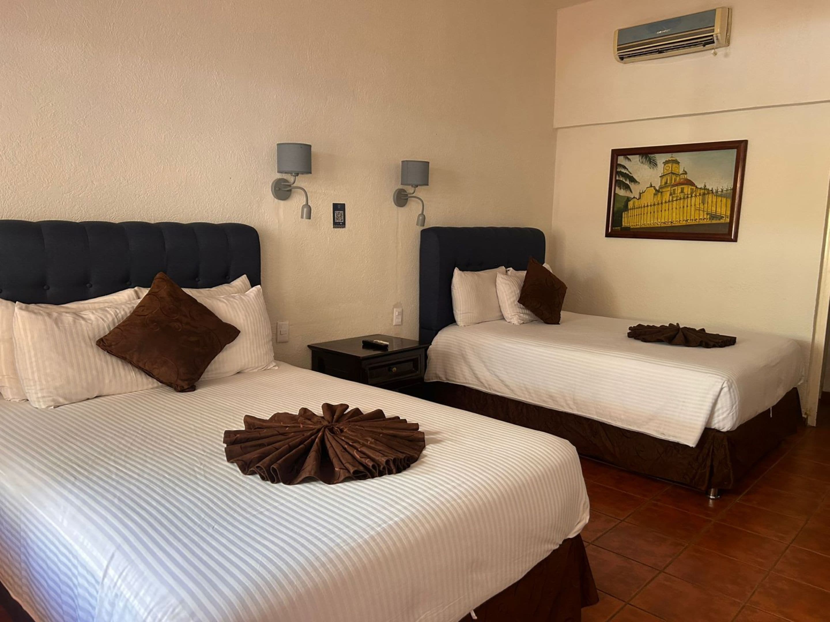 A hotel room with two beds, each with a headboard, white bedding, and a brown decorative pillow. There are two wall-mounted lamps and a painting of a building on the wall.
