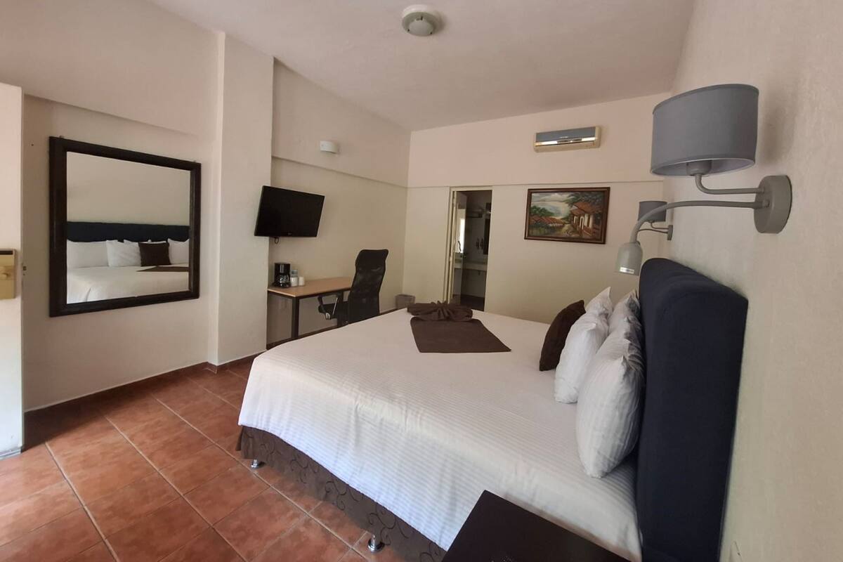 Ein Hotelzimmer mit einem großen Bett, einem Schreibtisch mit Stuhl, einem Fernseher, einem Bild an der Wand und einer Deckenleuchte.