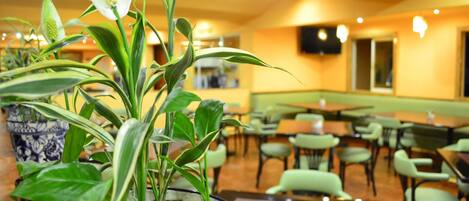 Interior de un restaurante con mesas y sillas, plantas en macetas y una pantalla de televisión en la pared.