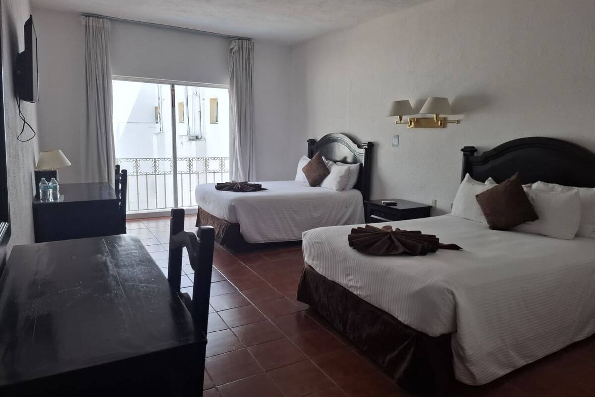 Ein Hotelzimmer mit zwei Betten, einem Schreibtisch, einem Stuhl, einem Fernseher und einem Balkon mit Vorhängen.