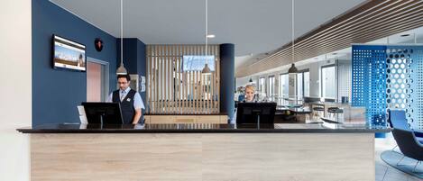 Un espace d’accueil d’hôtel au design moderne, comprenant un comptoir d’accueil, un écran numérique et un coin salon avec un mur bleu et des panneaux perforés.