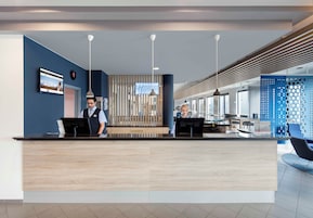 Zona de recepción de un hotel con diseño moderno, que incluye un mostrador de recepción, una pantalla digital y un área de descanso con pared azul y paneles perforados.
