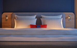 A neatly made bed with white linens and two pillows, a red accent pillow, and a blue tag in the center. - Holiday Inn Express München-Messe by IHG (Feldkirchen)
